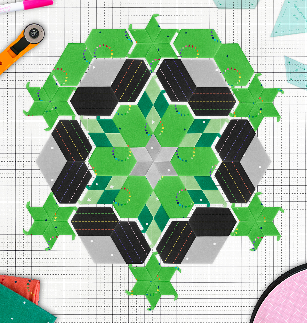 Green and gray English paper pieced block arranged on a white cutting board with fabric, templates and a rotary cutter scattered around the edge of the frame. 